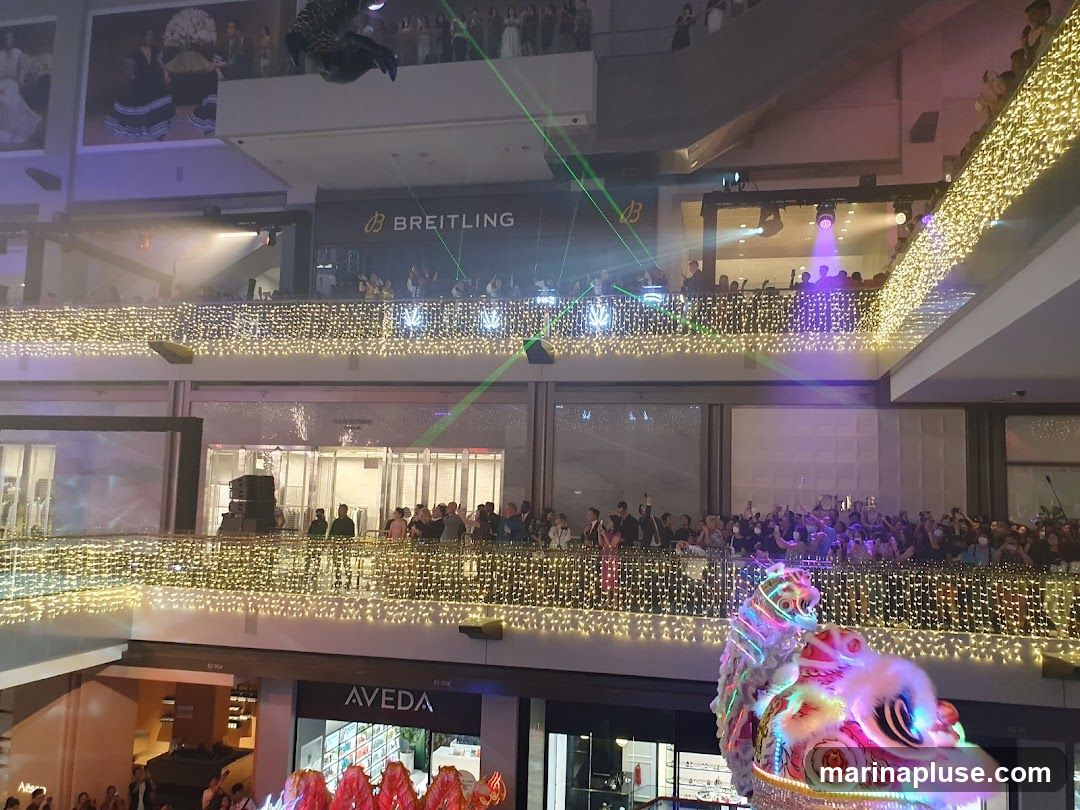 An indoor mall is decorated with numerous strings of golden fairy lights along its balconies. A performance, possibly a lion dance, is taking place in the foreground, with a colorful lion costume visible. People are gathered on multiple levels, watching the event. Laser lights in green and purple cut through the air. A Breitling store sign is visible on an upper level.