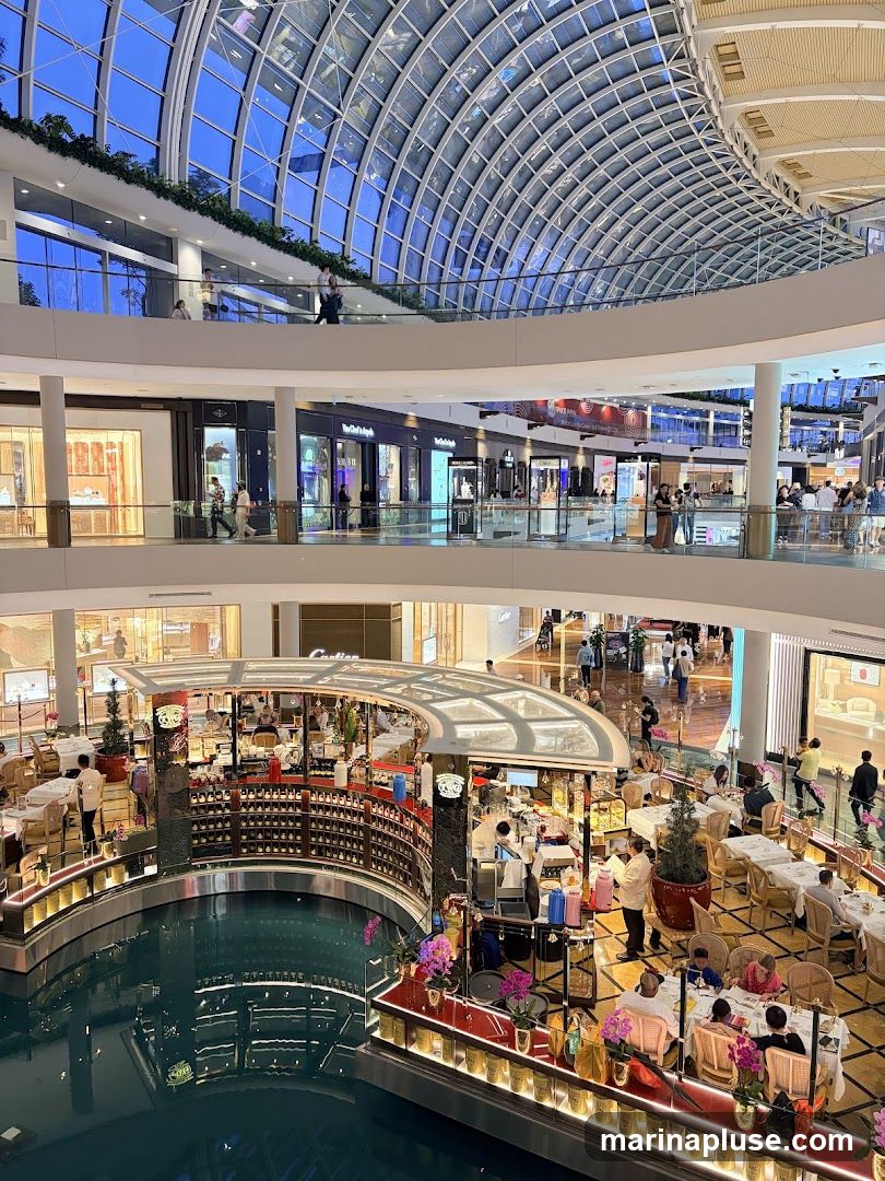 An interior view of a multi-level shopping mall with a large glass dome ceiling. A canal runs through the center of the mall, with a circular bar/restaurant area situated on it. People are seen dining at tables around the canal and browsing shops on the upper levels.