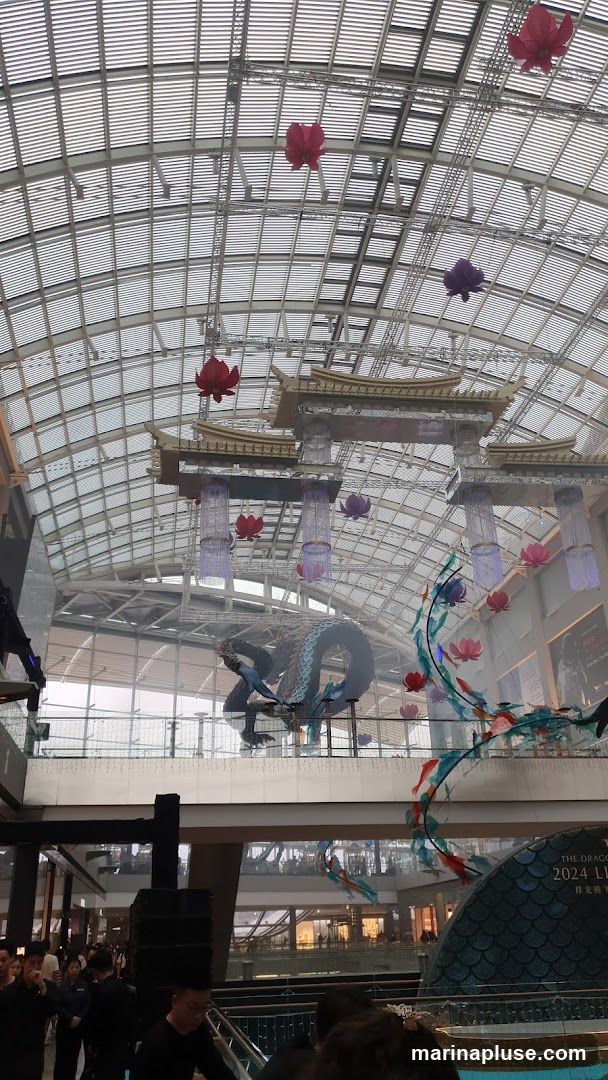 A large, blue dragon sculpture hangs from the ceiling of a modern shopping mall, surrounded by red and purple lotus flower decorations. The mall has a glass roof and multiple levels with shops visible in the background. A sign reads "THE DRAGON 2024 LUNAR NEW YEAR".
