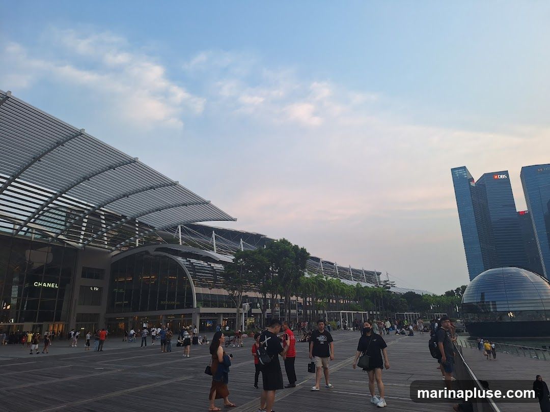 A wide shot of the Marina Bay Sands complex in Singapore. The iconic building with its distinctive sail-like roof is on the left, with the 'CHANEL' logo visible on its facade. In the background, the DBS and HSBC skyscrapers rise into the sky. A glass dome structure is visible to the right. People are walking on a wooden promenade in the foreground, enjoying the evening ambiance. The sky is a soft gradient of blue and pink, indicating either sunrise or sunset.