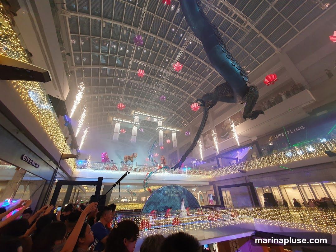An indoor atrium decorated for a festive event, featuring a large inflatable dragon suspended from the ceiling. Fireworks are set off at various levels, creating a dramatic effect. The scene is illuminated by numerous string lights and lanterns. A crowd of people is gathered, many holding up their phones to capture the spectacle. Storefronts, including GIVENCHY and BREITLING, are visible in the background, indicating this is likely a shopping mall. A stage with performers and a large circular backdrop is set up on a lower level.