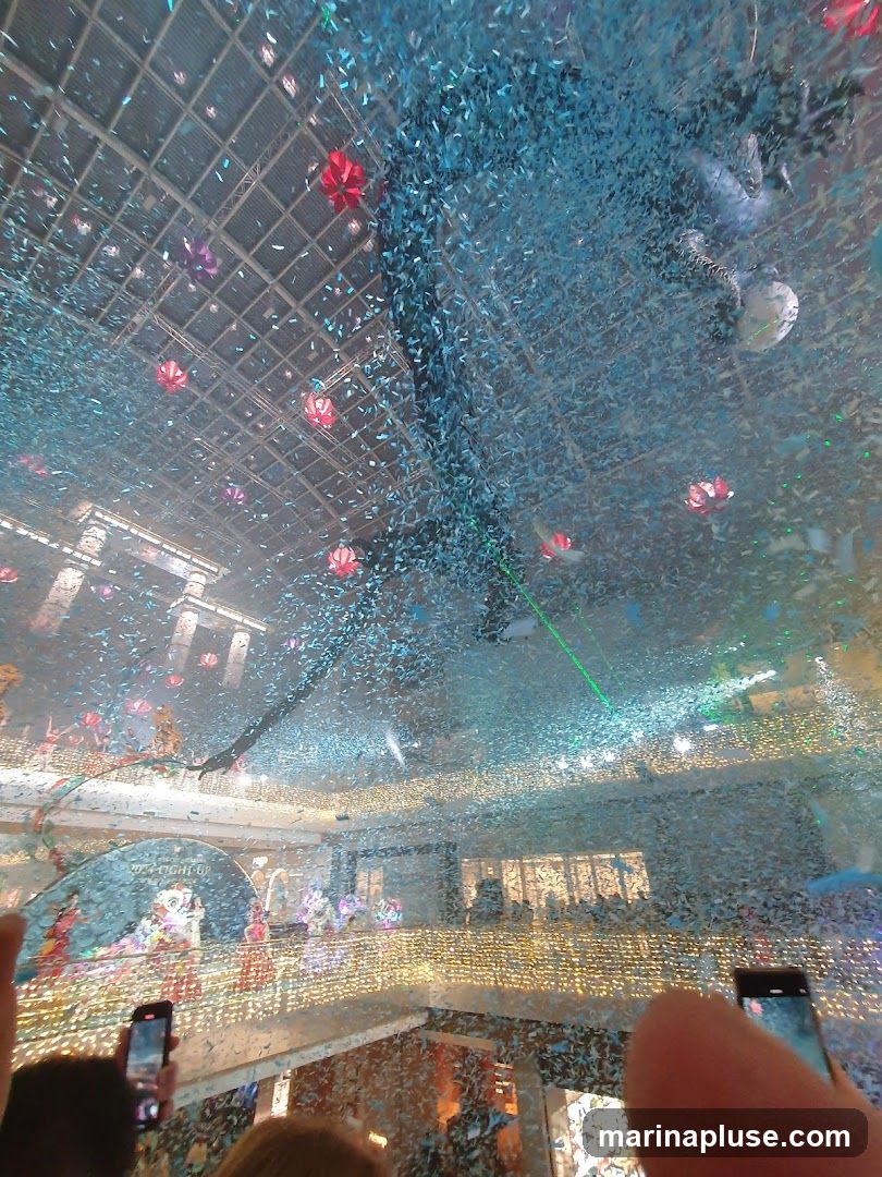 An indoor space, possibly a mall or event venue, is filled with a shower of blue and silver confetti. Red and purple decorative orbs hang from the ceiling. A stage or performance area is visible in the background with people watching. Several individuals are holding up smartphones to capture the event. Lights and a green laser beam add to the festive atmosphere.