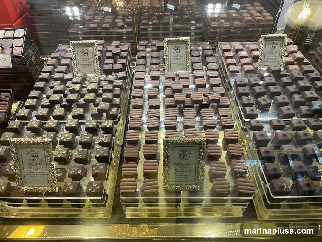A display case filled with various types of chocolates. The chocolates are arranged in rows on tiered stands. Several small framed signs with the TWG logo describe different chocolate flavors.