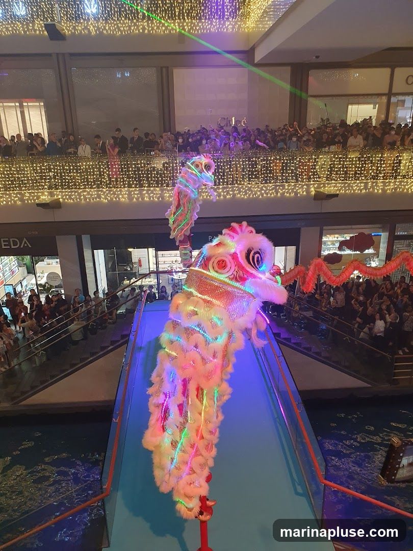 A vibrant lion dance performance is taking place in a multi-level indoor space, possibly a shopping mall. Two colorful lion costumes, adorned with LED lights, are the central focus. They are positioned on a raised platform above a body of water. A large crowd of spectators watches from balconies and walkways on different levels, illuminated by decorative string lights and lasers. Signs for businesses like "CHLOE" and "SEDA" are visible in the background, indicating a commercial setting.