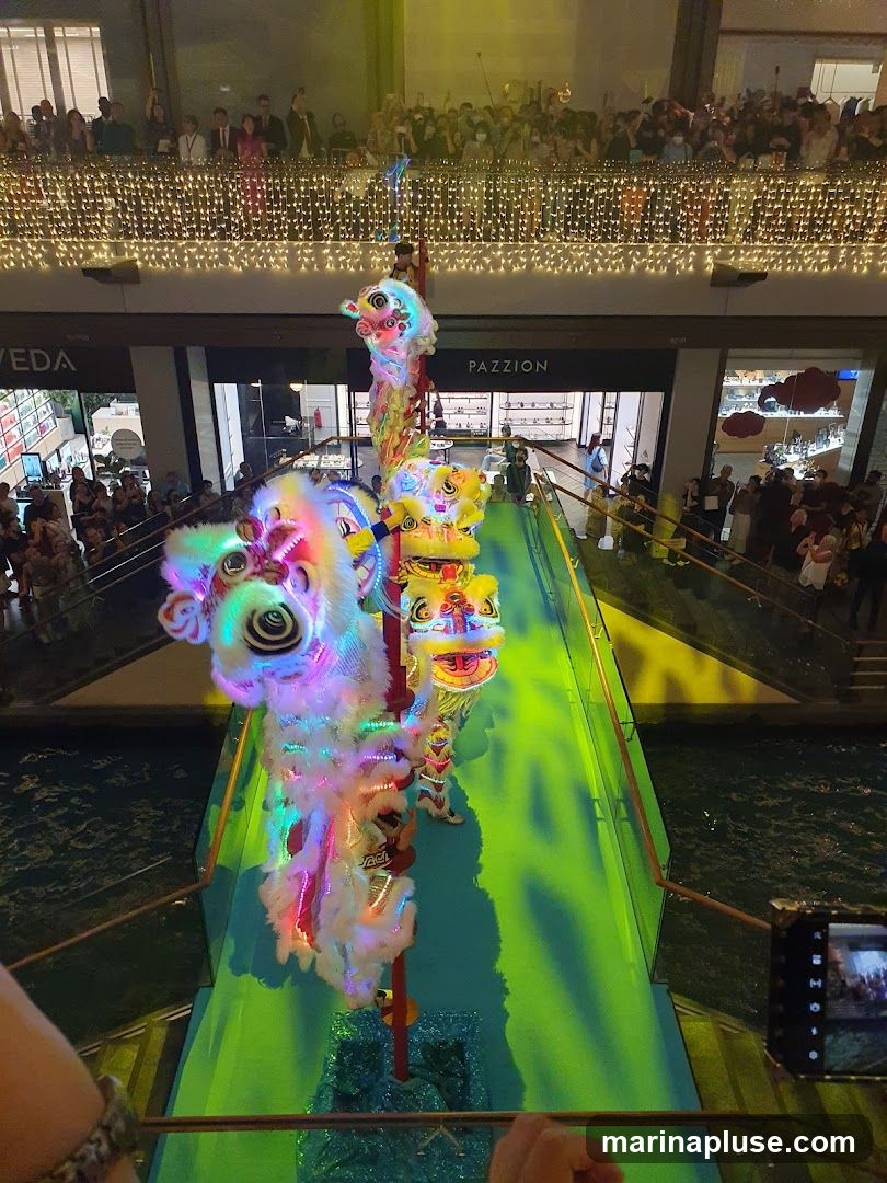 A group of performers in colorful lion costumes are dancing on a green illuminated walkway above water. A crowd of spectators watches from a balcony decorated with fairy lights. The scene appears to be an indoor shopping mall or entertainment venue.