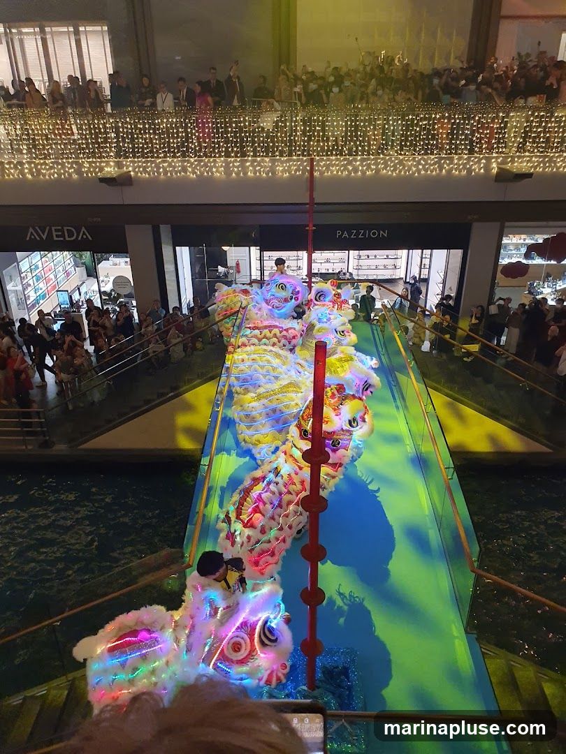 A vibrant lion dance performance is taking place on a blue and green walkway over water. The lions are colorful and illuminated with LED lights. A crowd of people watches from above and along the sides of the walkway. Two storefronts,