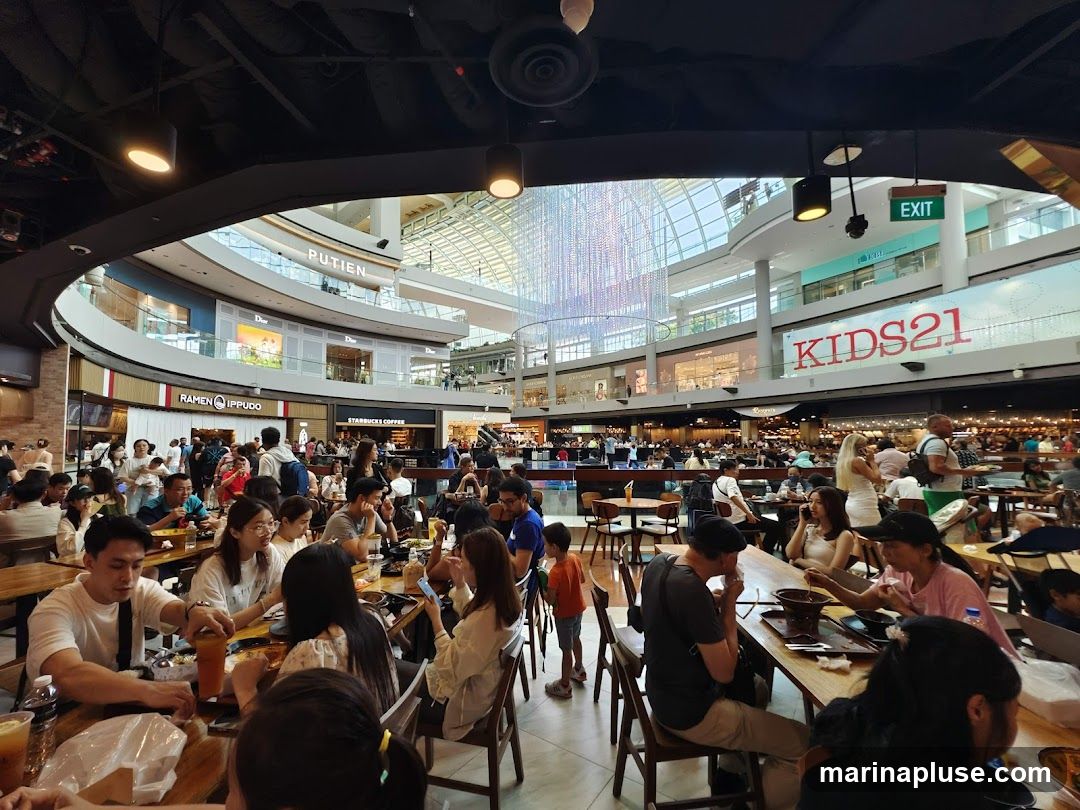 A bustling food court inside a modern shopping mall, filled with diners seated at various tables. The background shows multiple levels of shops, including 'PUTIEN', 'DIOR', 'RAMEN IPPUDO', 'STARBUCKS COFFEE', and 'KIDS21'. A large, decorative light installation hangs from the ceiling in the center of the atrium. People are seen eating, talking, and walking around.
