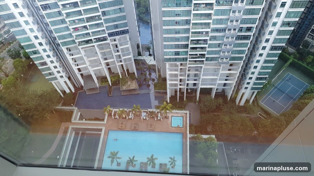 An aerial view of a modern apartment complex featuring multiple high-rise buildings, a swimming pool area with lounge chairs and umbrellas, a smaller jacuzzi-style pool, and a tennis court. Lush greenery surrounds the buildings and amenities. The image appears to be taken from a high vantage point, possibly a window of another building.
