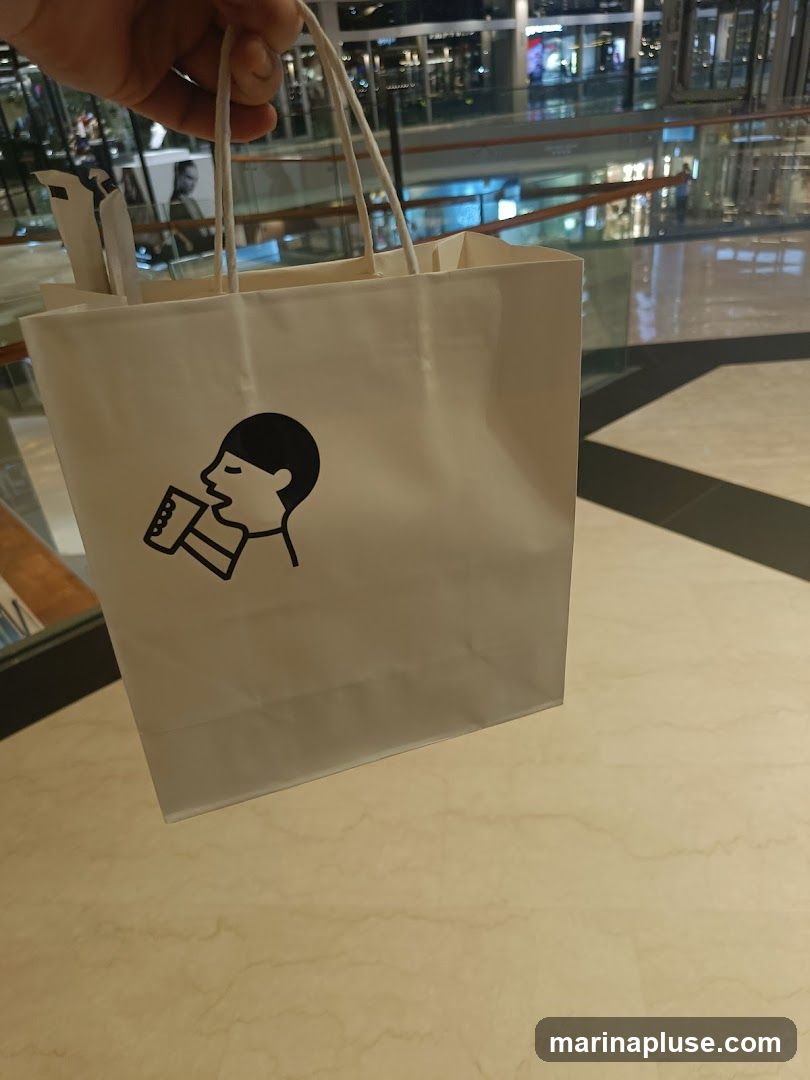 A white paper shopping bag with a black line drawing logo of a person drinking from a cup. The bag is held by a hand and is in the foreground, with a blurred background of a shopping mall interior.