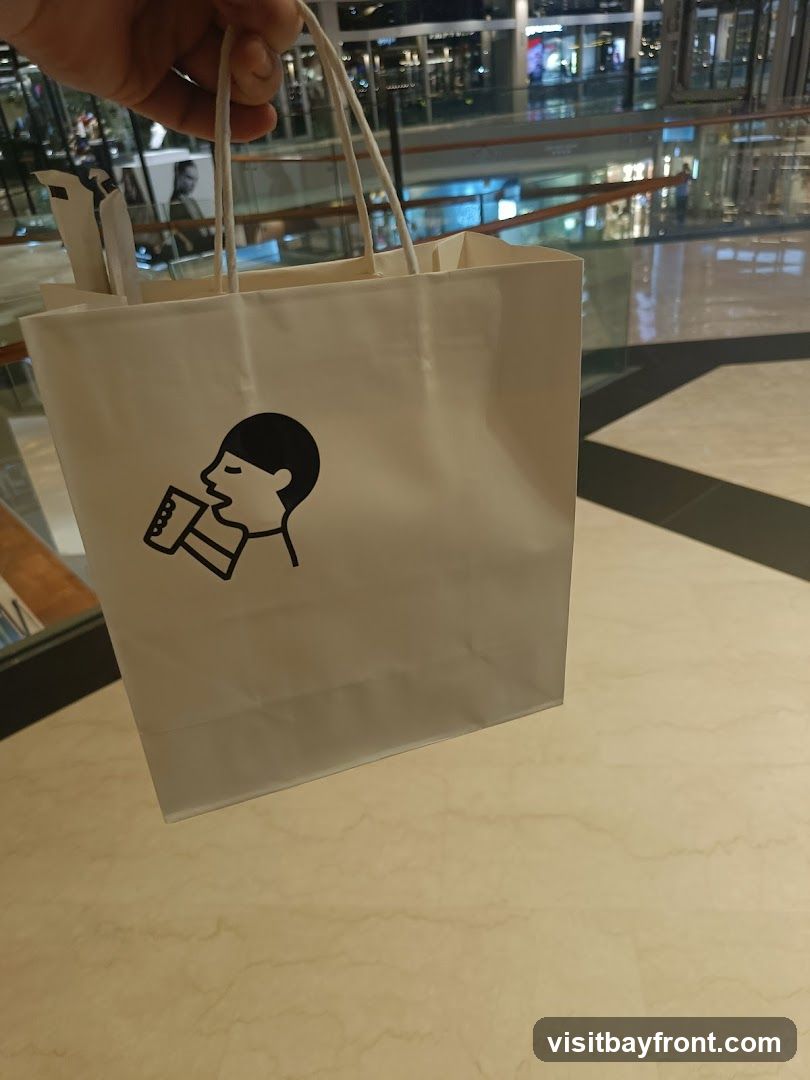 A white paper shopping bag with a black line drawing logo of a person drinking from a cup. The bag is held by a hand and is in the foreground, with a blurred background of a shopping mall interior.