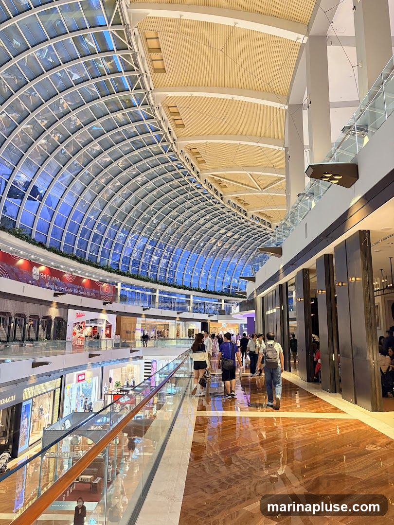 A wide shot of the interior of a large, modern shopping mall. The ceiling is a distinctive arched glass structure, allowing natural light to filter in. Below, a long walkway with polished wooden floors reflects the ambient light. People are seen walking along the walkway, browsing shops on multiple levels. Storefronts with various displays and signage are visible on either side. A banner with Japanese text and imagery is displayed on an upper level.