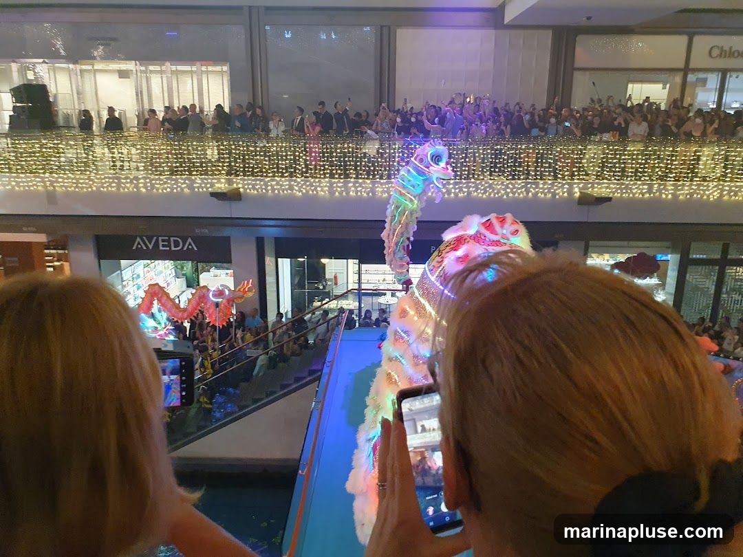 A lively dragon dance performance is taking place in a shopping mall, with a large crowd gathered to watch. The dragon is illuminated with colorful lights, and people are holding up their phones to capture the spectacle. The mall is decorated with festive lights, suggesting a celebration or event.