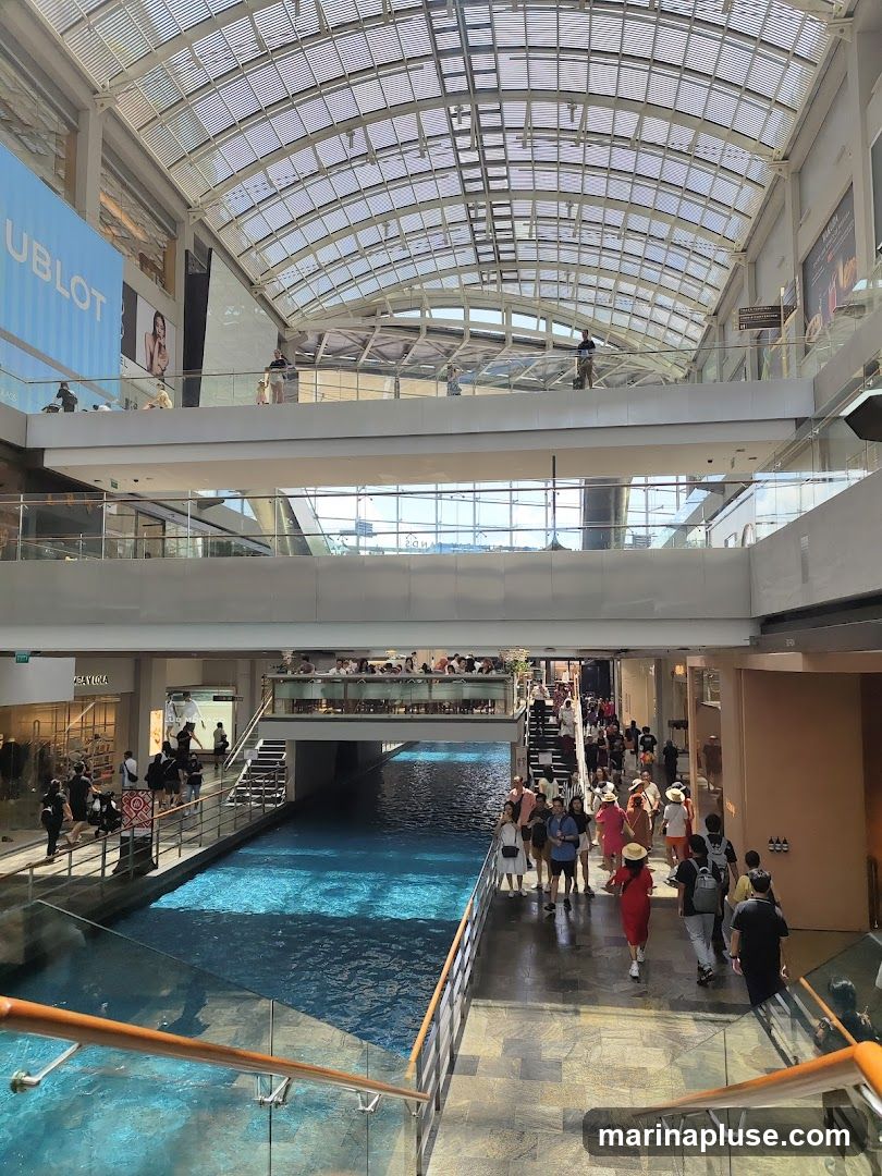 A view of a multi-level indoor shopping mall with a canal running through the center. People are walking on the walkways and on the stairs. Shops are visible on the sides, and a large HUBLOT advertisement is displayed on an upper level. The mall has a glass roof that allows natural light to enter.