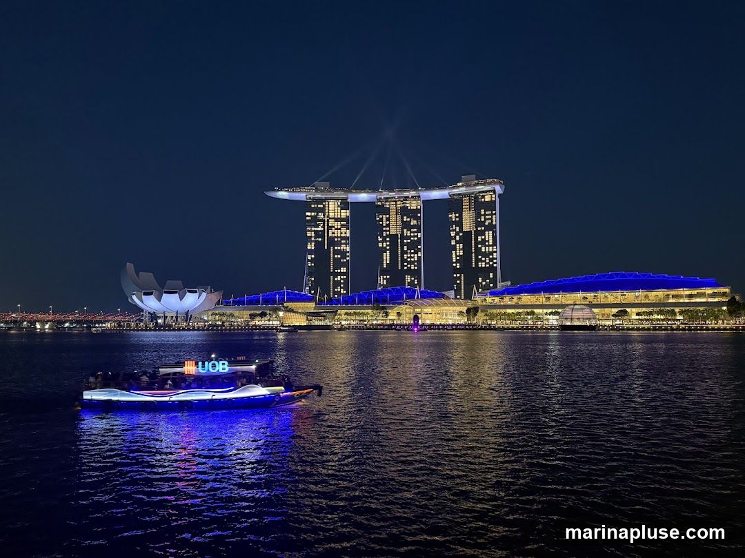 A nighttime view of the Marina Bay Sands complex in Singapore, with its iconic three towers and the ArtScience Museum illuminated. A boat with the UOB logo is visible in the foreground on the water, lit with blue and white lights.