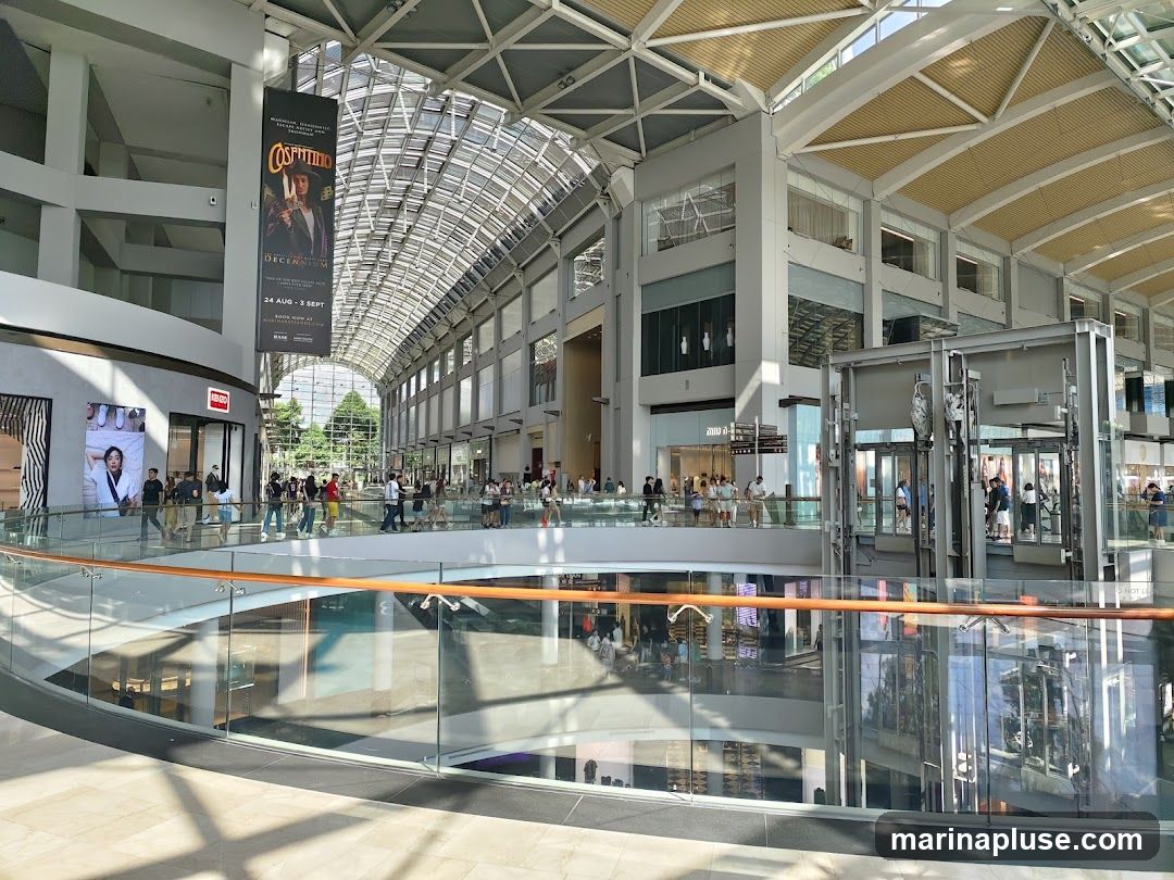 A wide shot of the interior of a large, modern shopping mall. The architecture features a high, vaulted glass ceiling with a metal support structure. Multiple levels of shops and walkways are visible, with glass railings and escalators. People are seen browsing and walking around. A large banner advertising 'Costumes Decorum' hangs from the ceiling.