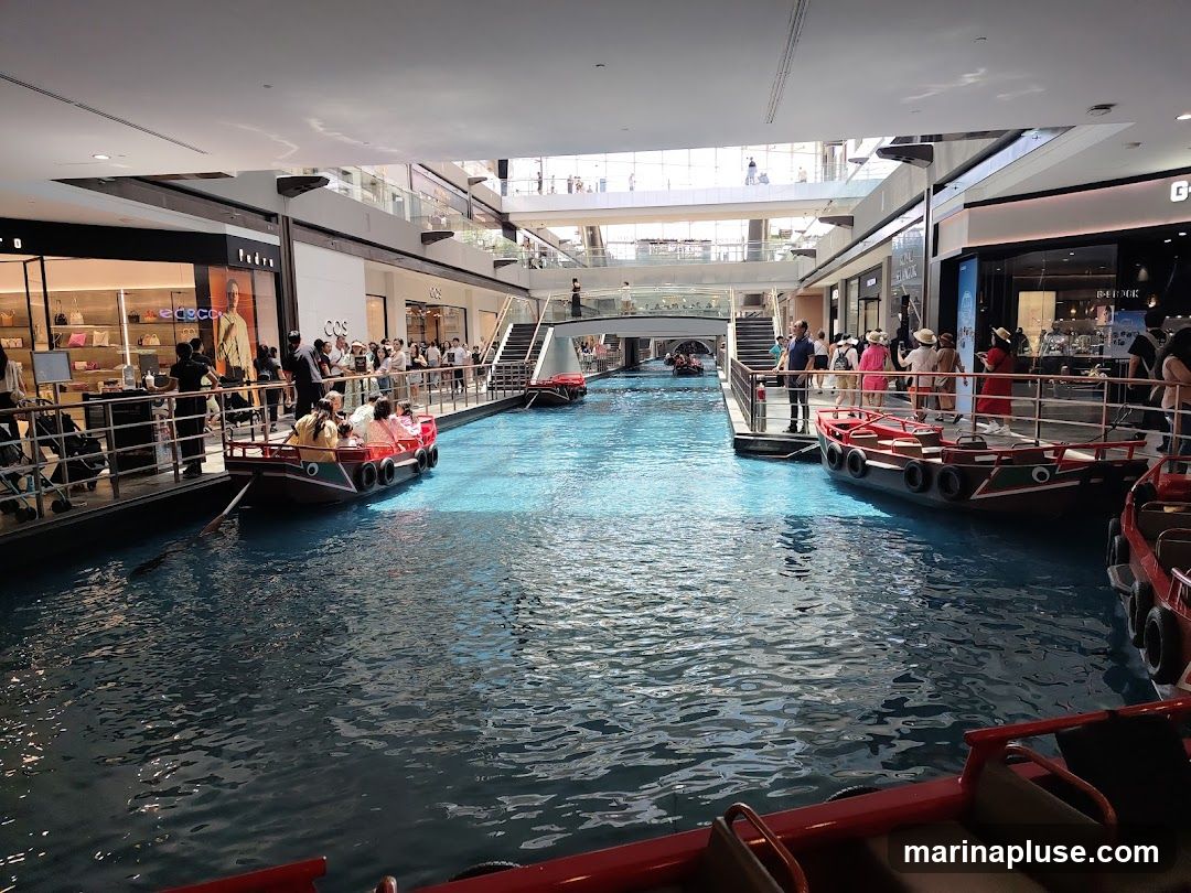 An indoor canal with several gondola-style boats carrying passengers. The canal is surrounded by a multi-level shopping mall with various storefronts visible. People are seen walking along the canal's edge and riding in the boats. The architecture suggests a modern, upscale shopping environment.