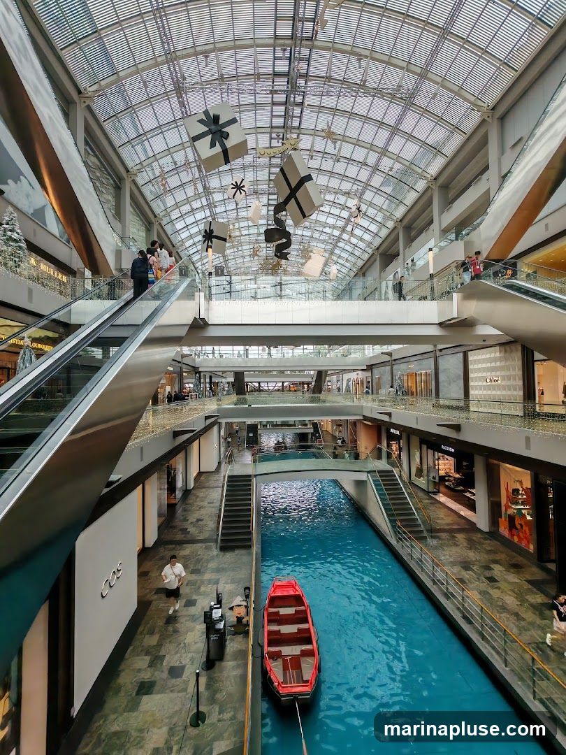 An interior view of a large, modern shopping mall with a glass ceiling. A bright blue canal runs through the center of the mall, with a small red boat docked. Escalators and walkways are visible on multiple levels, with various shops lining the sides. Decorative gift boxes and stars hang from the ceiling, suggesting a festive atmosphere. People are seen shopping and walking around.
