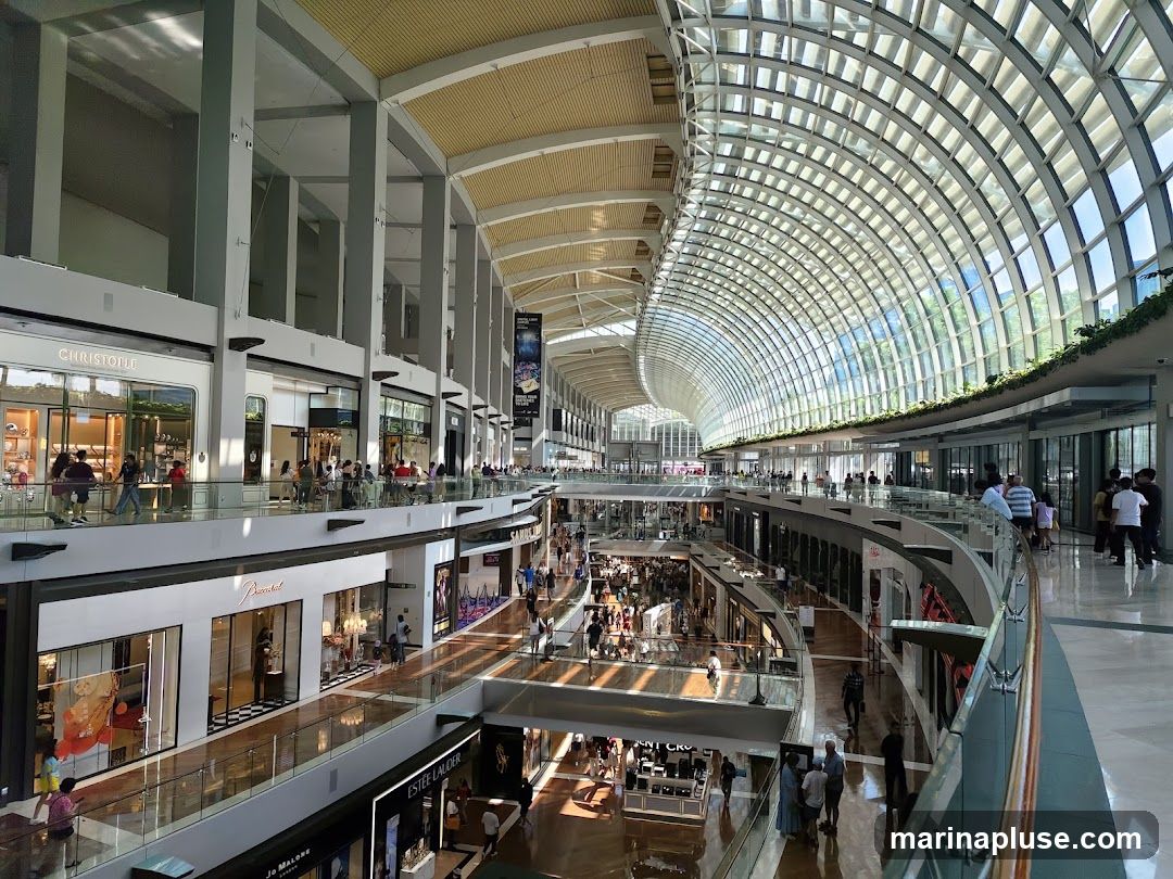 An interior view of a multi-level shopping mall with a distinctive curved glass roof. Shops are visible on various levels, with escalators and walkways connecting them. Many shoppers are present, browsing the stores and moving around the mall. The architecture features large pillars and a bright, airy atmosphere due to the natural light filtering through the roof.