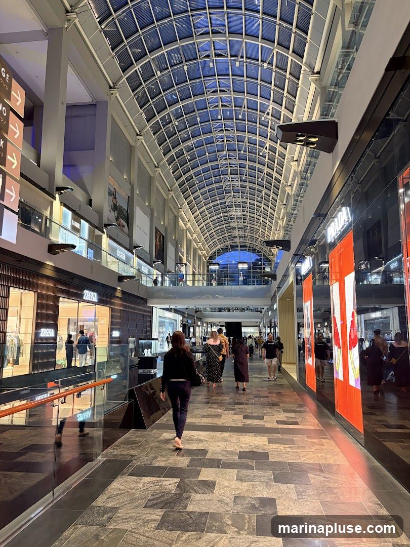 A wide, modern indoor shopping mall corridor with a high, arched glass ceiling. The corridor is lined with various shops on both sides. People are seen walking along the corridor. On the left, there are directional signs with Japanese characters. On the right, large digital screens display vibrant advertisements. The floor is paved with large, dark tiles arranged in a geometric pattern.