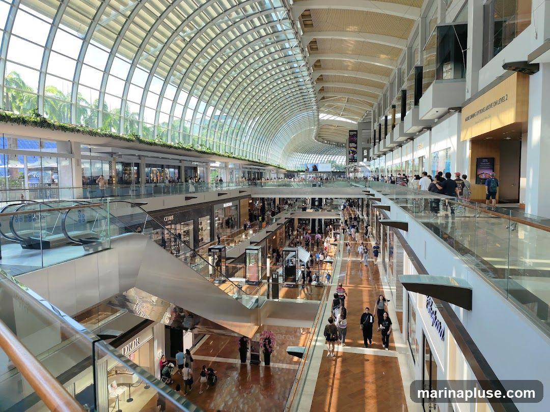 The image shows the interior of a spacious, multi-level shopping mall with a distinctive arched glass roof. Sunlight streams through the roof, illuminating the polished floors and the various shops lining the walkways. Escalators and walkways connect different levels, and numerous shoppers are seen browsing and walking around. Storefronts for luxury brands like Dior and Guess are visible. Greenery is incorporated into the mall's design, with plants adorning the upper levels.