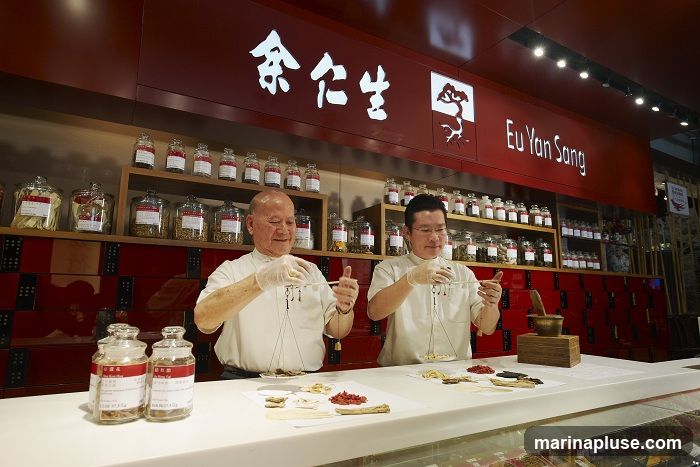 Two men in white coats are weighing herbs on traditional scales behind a counter in a shop. The shop has shelves filled with jars of herbs and a prominent sign with Chinese characters and "Eu Yan Sang" written on it.