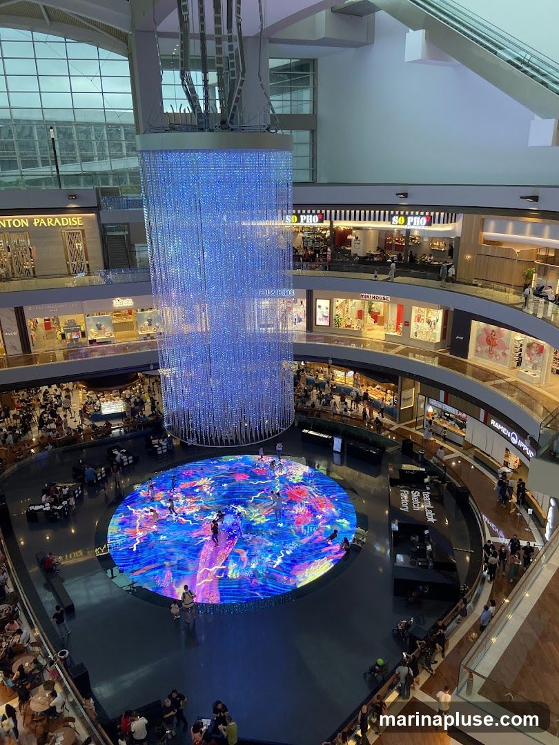 An overhead view of a bustling shopping mall interior. A large, multi-story digital art installation with vibrant, swirling colors is displayed in the center of the atrium, with people interacting with it. Above the installation, a massive crystal chandelier hangs, casting a blue light. Surrounding the atrium are multiple levels of shops and restaurants, with shoppers browsing and dining. Visible shop names include 'SO PHO', 'FILA KIDS', 'MIKIHOUSE', 'RATIONALE', and 'RAMEN IPPUDO'.