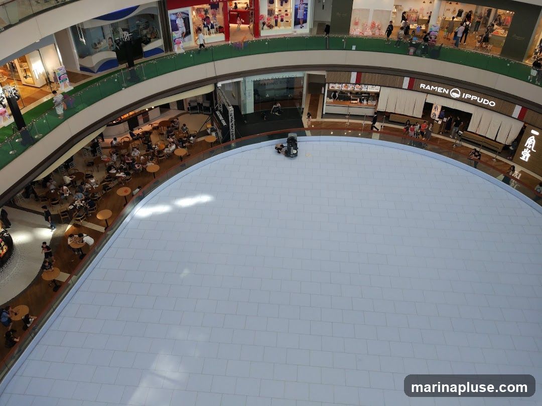 An aerial view of an indoor ice rink in a multi-level shopping mall. People are seen skating on the rink and dining at tables surrounding it. Various shops and escalators are visible on the upper levels. A ramen restaurant with visible signage is also present.