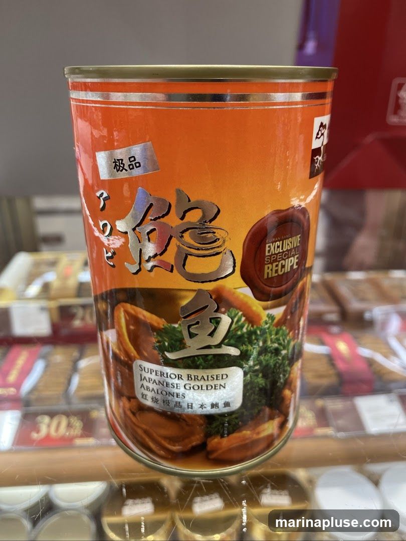 A close-up of a can of Superior Braised Japanese Golden Abalones. The can is orange with prominent Chinese characters and an image of abalones and greens. A seal indicates an "Exclusive Special Recipe". In the background, shelves with other products and price tags are visible.
