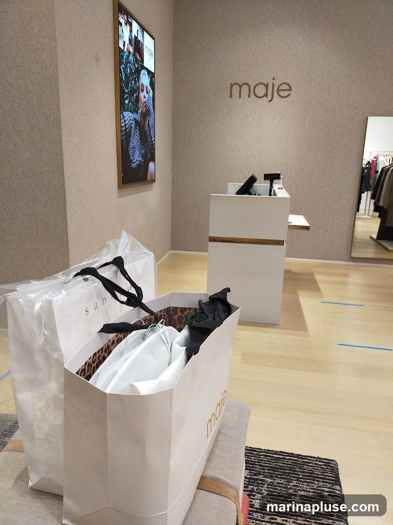 A retail store interior with shopping bags in the foreground. A large white shopping bag with the logo 'maje' is filled with clothing items. Another white shopping bag with the logo 'sandro' is partially visible to the left. A digital screen displaying fashion imagery is mounted on the wall. The store features a checkout counter and a mirror reflecting clothing racks. The walls are textured beige, and the floor is light wood. Blue tape lines are visible on the floor, possibly indicating social distancing markers.