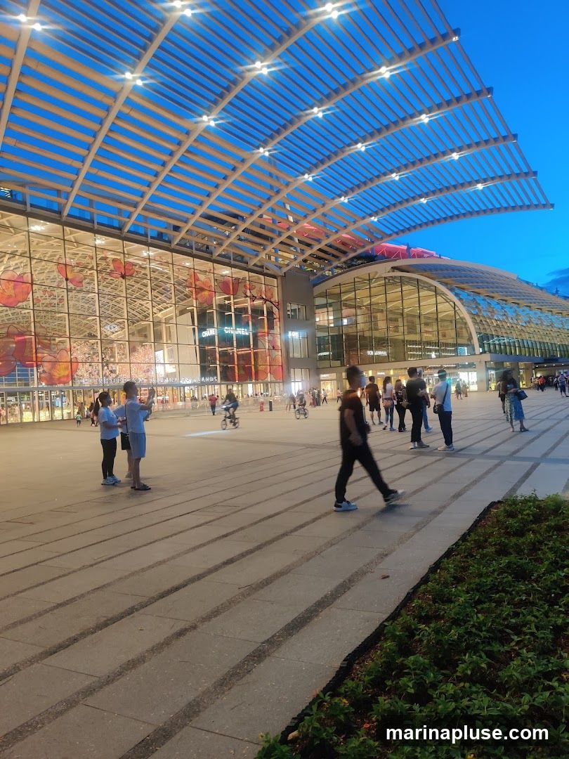 A wide shot of a modern building with a large, curved glass facade and a slatted roof structure. The building is illuminated by lights, and the sky is a deep blue, indicating dusk. People are walking in the plaza in front of the building, and some are on bicycles. The facade features large floral graphics and visible signage for 'CHANEL'.