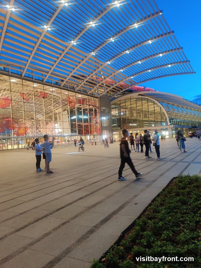 A wide shot of a modern building with a large, curved glass facade and a slatted roof structure. The building is illuminated by lights, and the sky is a deep blue, indicating dusk. People are walking in the plaza in front of the building, and some are on bicycles. The facade features large floral graphics and visible signage for 'CHANEL'.