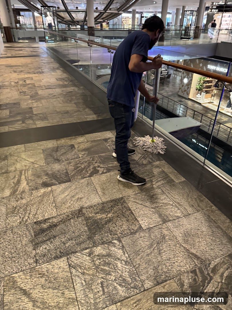 A man in a blue t-shirt and jeans is mopping the floor of a modern shopping mall. He is standing on a walkway with large, patterned tiles. In the background, there are shops with glass storefronts and escalators. One visible sign reads "TORY BURCH" on a shop facade.