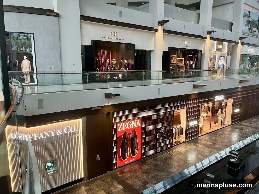 An interior view of a multi-level shopping mall featuring several high-end retail stores. On the lower level, Tiffany & Co. is visible with its illuminated signage and display window. Adjacent to it is a Zegna store, showcasing clothing on mannequins and a large advertisement for Zegna shoes. On the upper level, stores such as Carolina Herrera and La Mer are present, with mannequins displaying fashion items in their windows. The mall has a modern architectural design with glass railings and escalators.