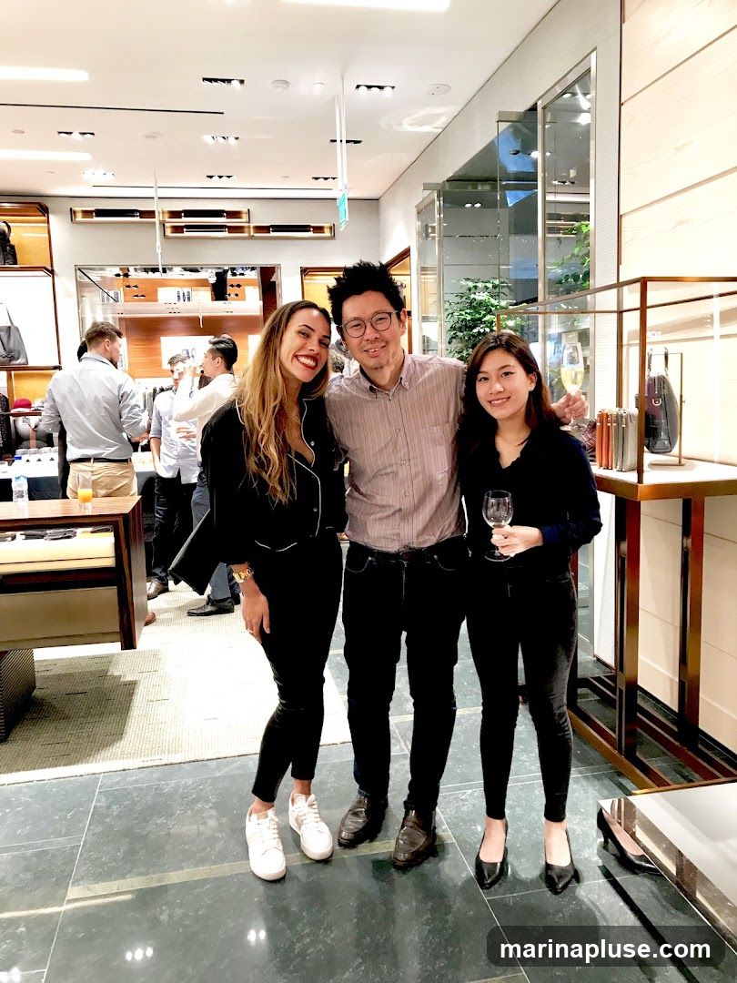 Three people, two women and one man, are posing for a photo in what appears to be a retail store. The woman on the left is wearing a black top and black pants with white sneakers. The man in the middle is wearing a striped button-down shirt and dark pants with dress shoes. The woman on the right is wearing a black top and black pants with black heels, holding a wine glass. The background shows shelves stocked with merchandise, possibly handbags and accessories, and other shoppers.