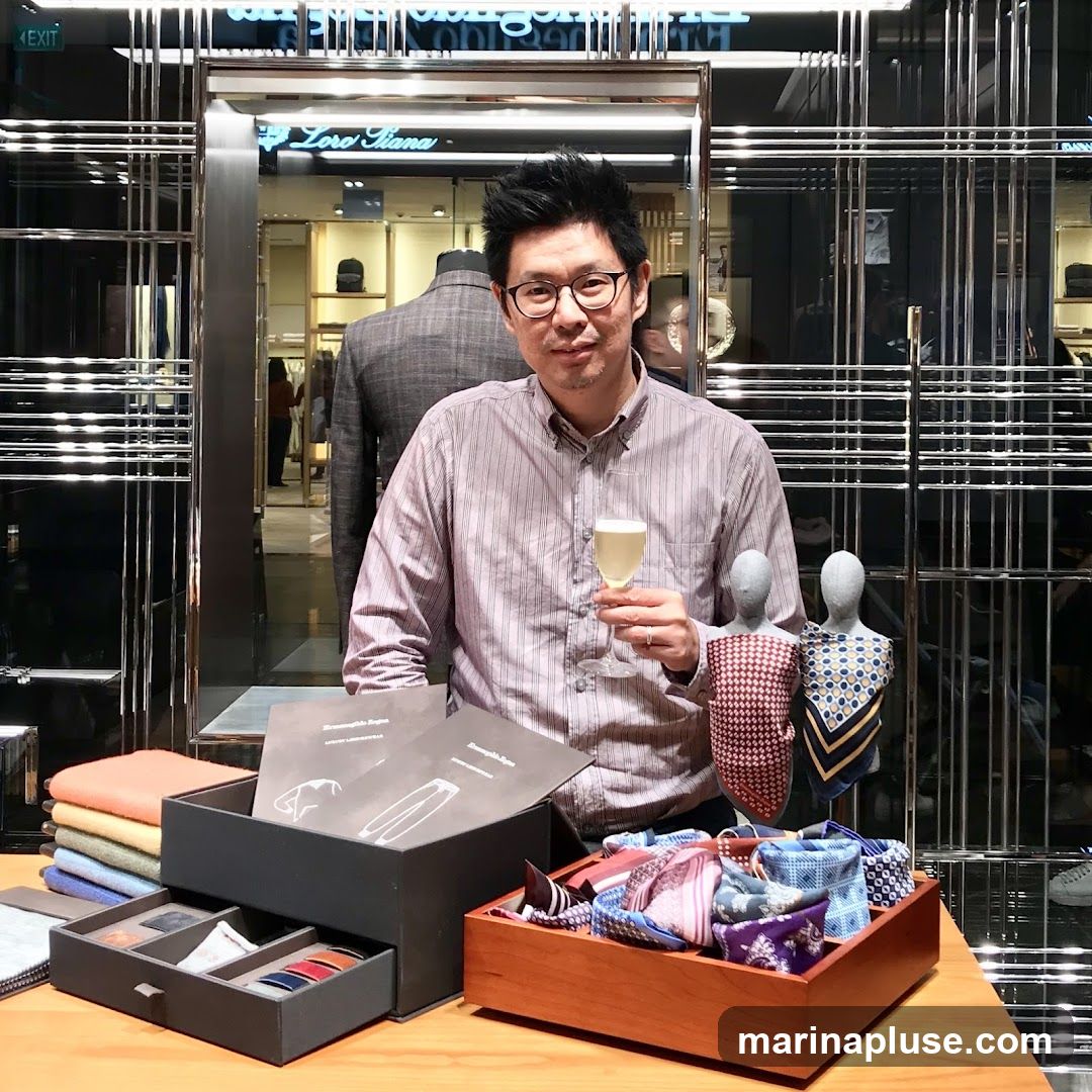 A man in a striped shirt and glasses holds a glass of champagne while standing in front of a display of men's accessories, including ties and pocket squares. Behind him, a mannequin is dressed in a suit, and the store's signage, "Loro Piana," is visible. The store appears to be a high-end boutique with a modern, sleek interior design.
