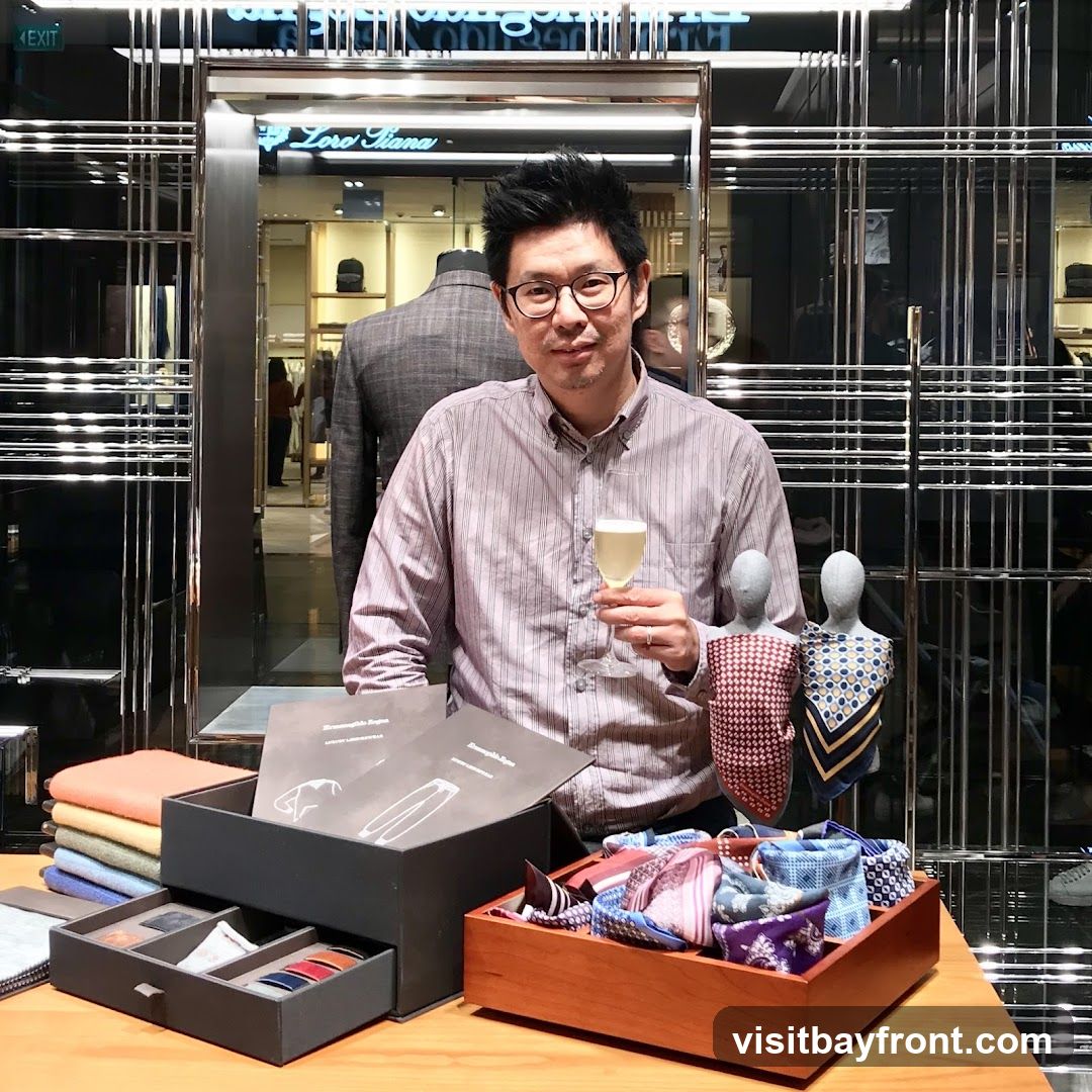 A man in a striped shirt and glasses holds a glass of champagne while standing in front of a display of men's accessories, including ties and pocket squares. Behind him, a mannequin is dressed in a suit, and the store's signage, "Loro Piana," is visible. The store appears to be a high-end boutique with a modern, sleek interior design.