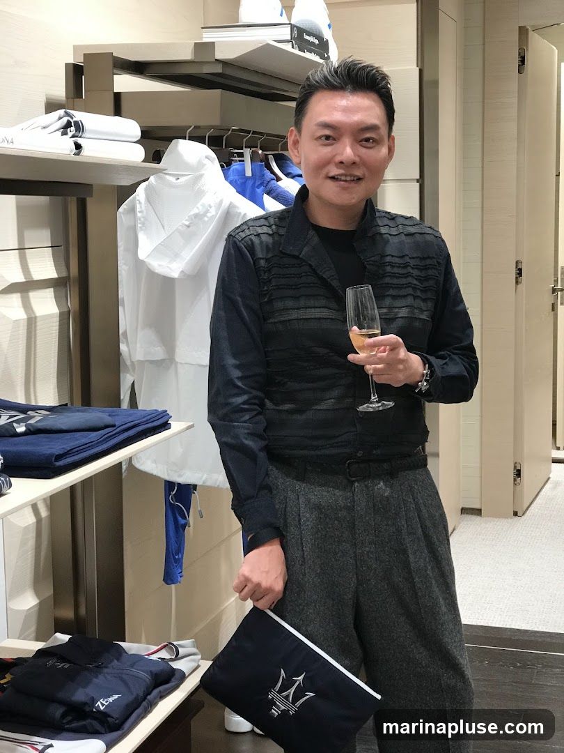 A man stands in a store, holding a glass of champagne and a dark blue clutch bag with a silver trident logo. He is wearing a dark, textured shirt and grey trousers. Behind him, clothing is displayed on shelves and racks, including folded shirts and jackets. The store appears to be a high-end boutique.