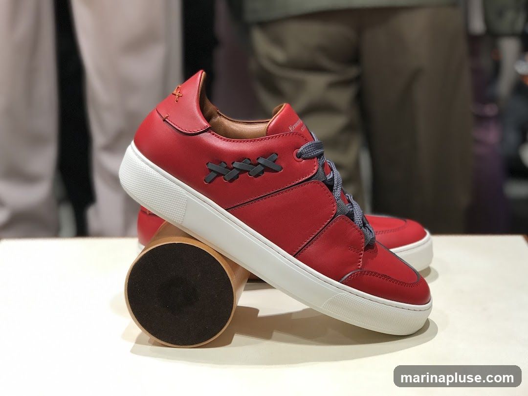 A pair of red leather sneakers with grey laces and white soles are displayed on a white surface, supported by a wooden cylinder. The sneakers have a unique criss-cross stitching detail on the sides. In the background, blurred figures in casual attire are visible, suggesting a retail environment.