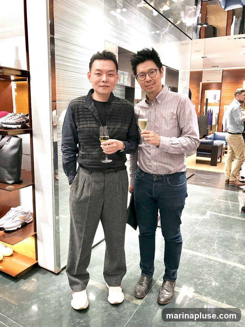 Two men are standing in a well-lit store, holding champagne glasses. The man on the left is wearing a dark, textured long-sleeved shirt and grey pleated trousers. The man on the right is wearing a striped button-down shirt and dark jeans. In the background, shelves display shoes and bags, and other shoppers can be seen browsing. The store appears to be a high-end retail establishment.