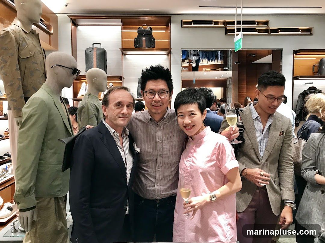 A group of people are posing for a photo inside a well-lit retail store. The store appears to be a high-end fashion boutique, with mannequins displaying clothing and accessories like bags and shoes. The individuals in the foreground are smiling and holding glasses of champagne, suggesting a celebratory event or product launch. The background shows shelves stocked with merchandise and an exit sign.
