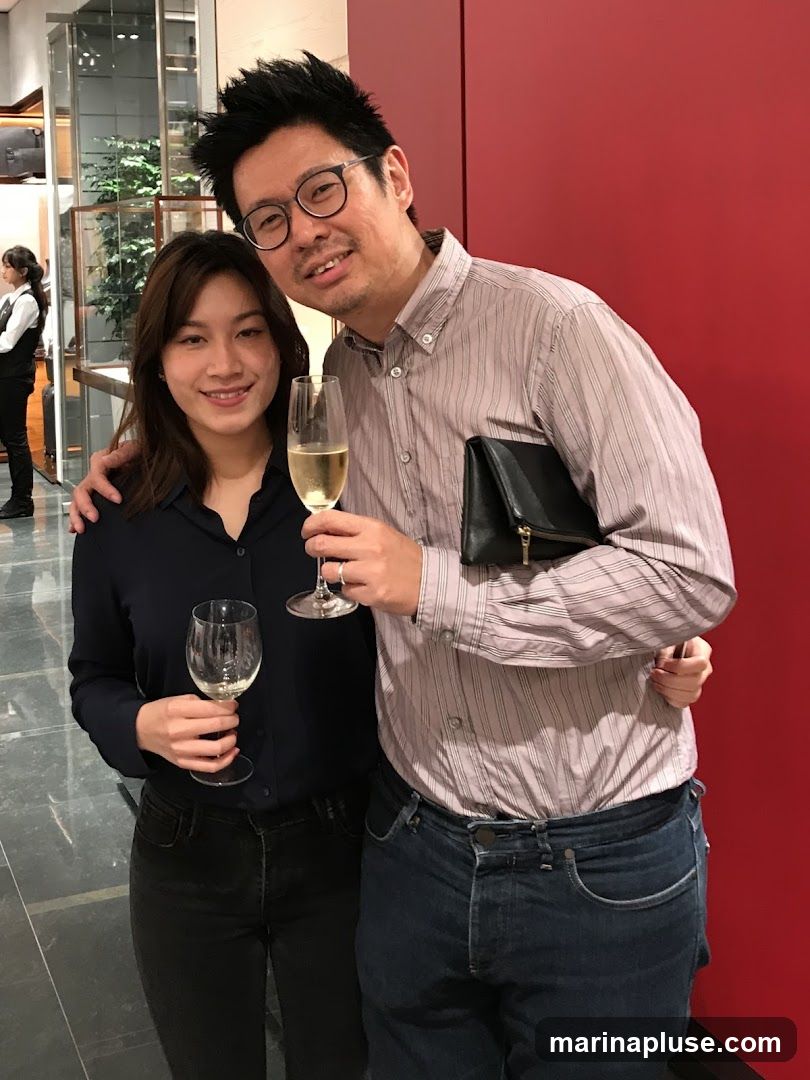 A man and a woman are posing for a photo, both holding champagne glasses. The man is wearing a striped button-down shirt and jeans, and the woman is wearing a dark blue collared shirt and black jeans. They are standing in front of a red wall, with a display case and a person in uniform visible in the background.