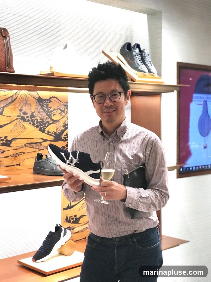 A man in a striped shirt and jeans holds a black and white sneaker in his left hand and a glass of champagne in his right. He is standing in front of shelves displaying various shoes and bags. The background shows a modern retail interior with minimalist displays.