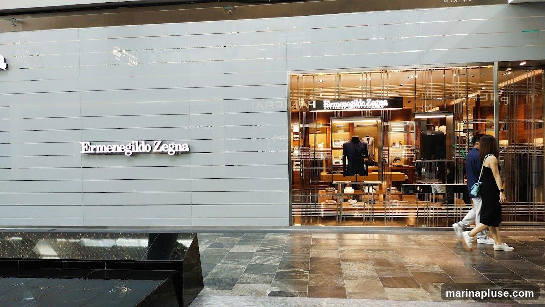 The exterior of an Ermenegildo Zegna store. The storefront features a large glass facade with a metallic grid design. Inside, mannequins display suits and other clothing items. The store's name is visible on the facade and also as a prominent sign on the left side of the building. Two people are walking past the store.