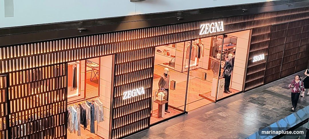 The exterior of a Zegna store with illuminated signage. The storefront features a modern design with vertical wooden slats and large glass windows displaying clothing on mannequins and racks.
