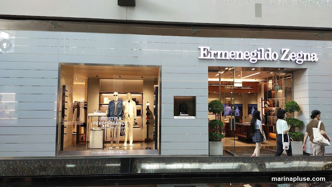 The exterior of a luxury clothing store, Ermenegildo Zegna, with a large display window showcasing mannequins dressed in stylish outfits. The store features a modern facade with horizontal paneling and prominent signage. Inside, racks of clothing and accessories are visible, along with decorative plants. Several people are walking past the storefront.
