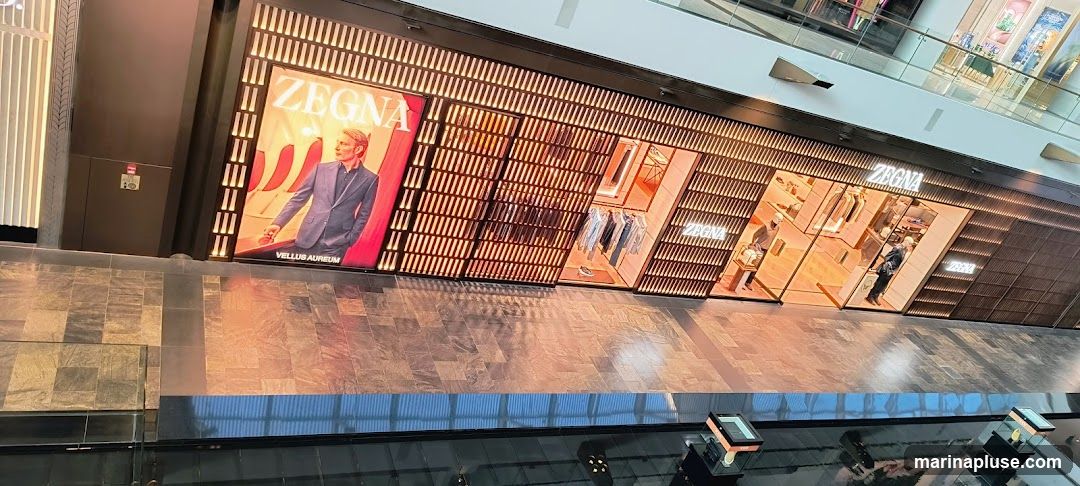 The exterior of a Zegna store with a large display featuring a man in a suit and the Zegna logo. The store has a modern design with wooden slatted panels and illuminated displays showcasing clothing and accessories.
