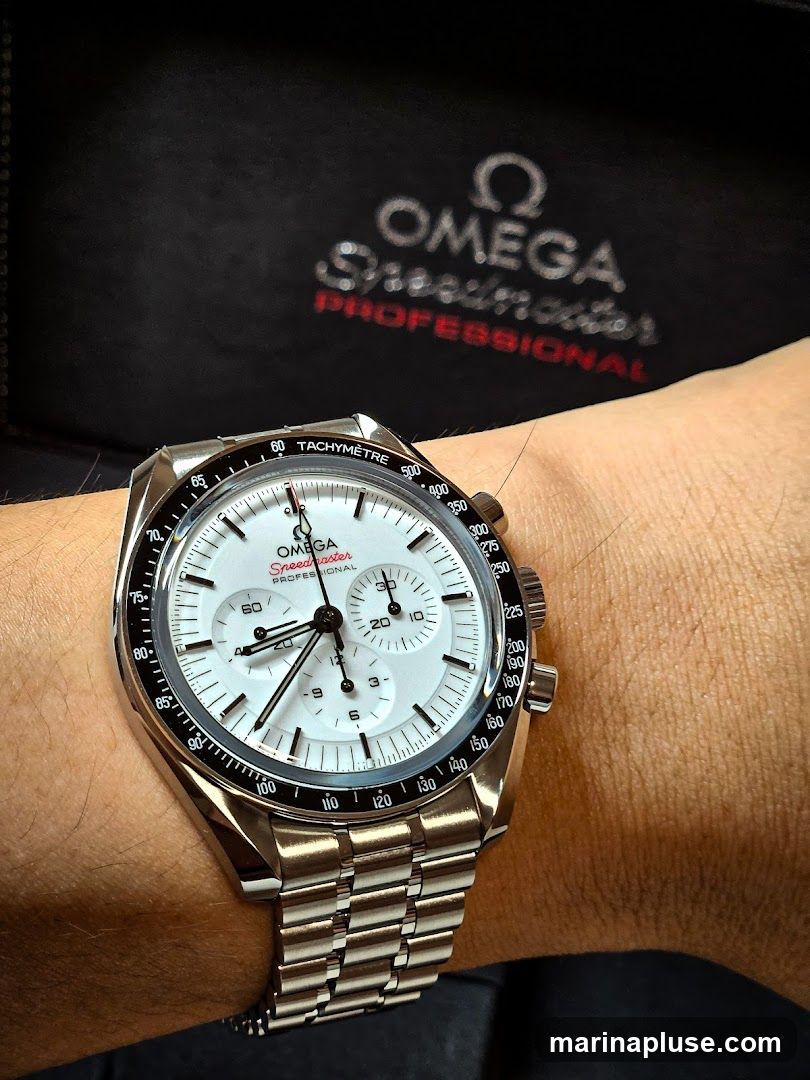 A close-up shot of an Omega Speedmaster Professional watch on a person's wrist. The watch has a white dial with silver hands and markers, and a tachymeter bezel. In the background, the Omega logo and "Speedmaster Professional" text are visible on a black surface.