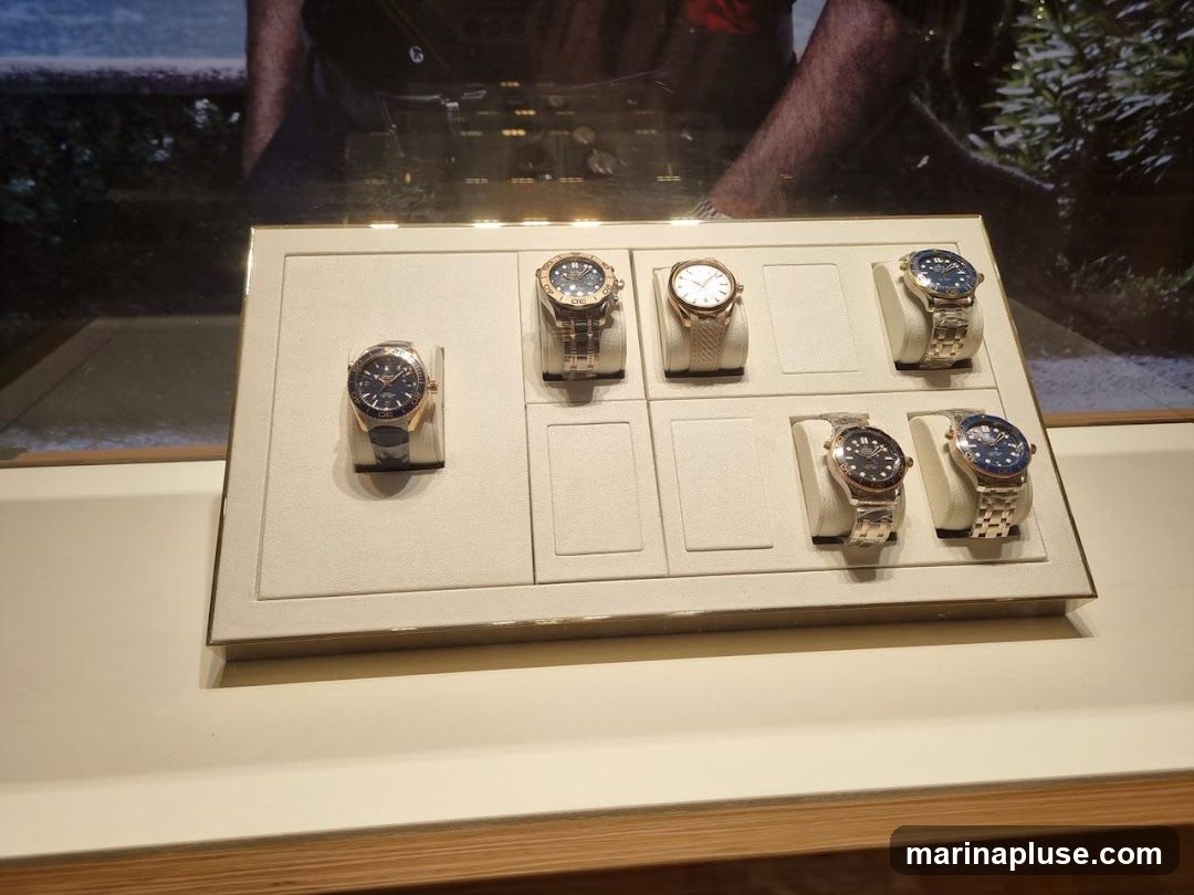 A display case shows six luxury watches. The watches are arranged in two rows of three. The top row has three watches, and the bottom row has three watches. The watches are on white stands. The background shows a blurred image of a person and a natural scene.