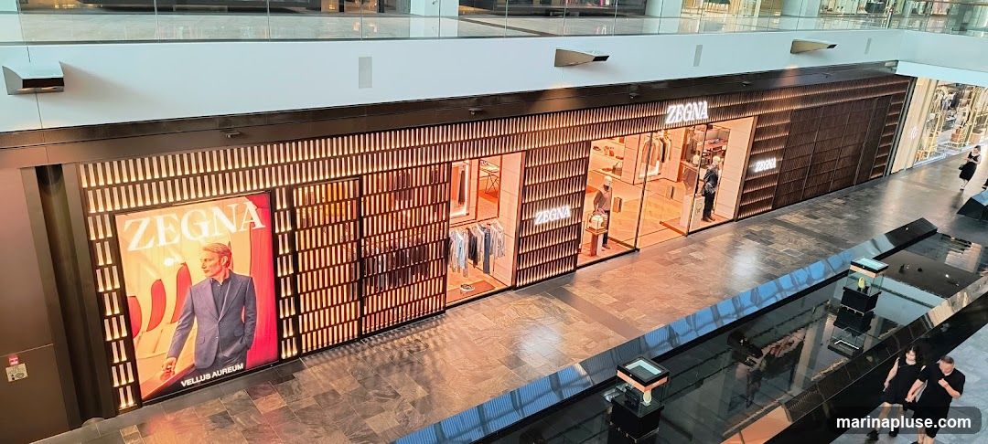 A wide shot of a Zegna store in a modern mall. The storefront features a large illuminated advertisement with a man in a suit, and the Zegna logo prominently displayed. The store interior is visible through large glass windows, showcasing clothing racks with men's apparel and mannequins dressed in suits. The facade is decorated with vertical wooden slats and warm lighting.