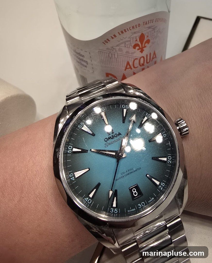 A close-up shot of an Omega Seamaster watch on a person's wrist. The watch has a blue dial with silver hands and hour markers. The date window shows the number 8. In the background, a bottle with a label that reads "ACQUA" and "FOR AN ENRICHED TASTE EXPERIEN" is partially visible.